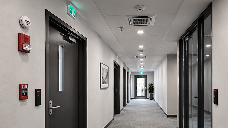 Modern commercial corridor with integrated fire alarm components including smoke detectors, strobe horn, pull station, sprinkler head, and emergency exit signage.