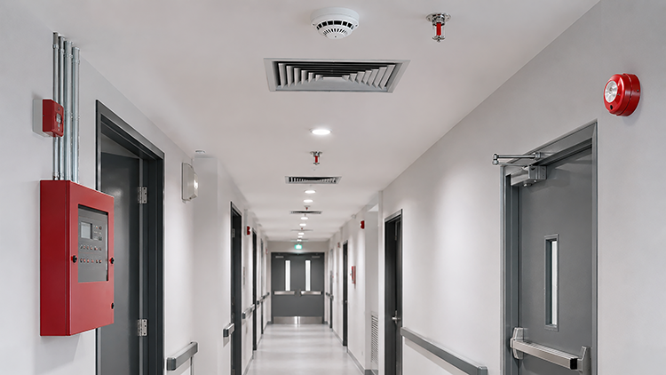 Commercial corridor with ceiling-mounted smoke detectors, fire sprinkler heads, a red alarm strobe, and a wall-mounted fire alarm panel showing integrated life safety components throughout the building.